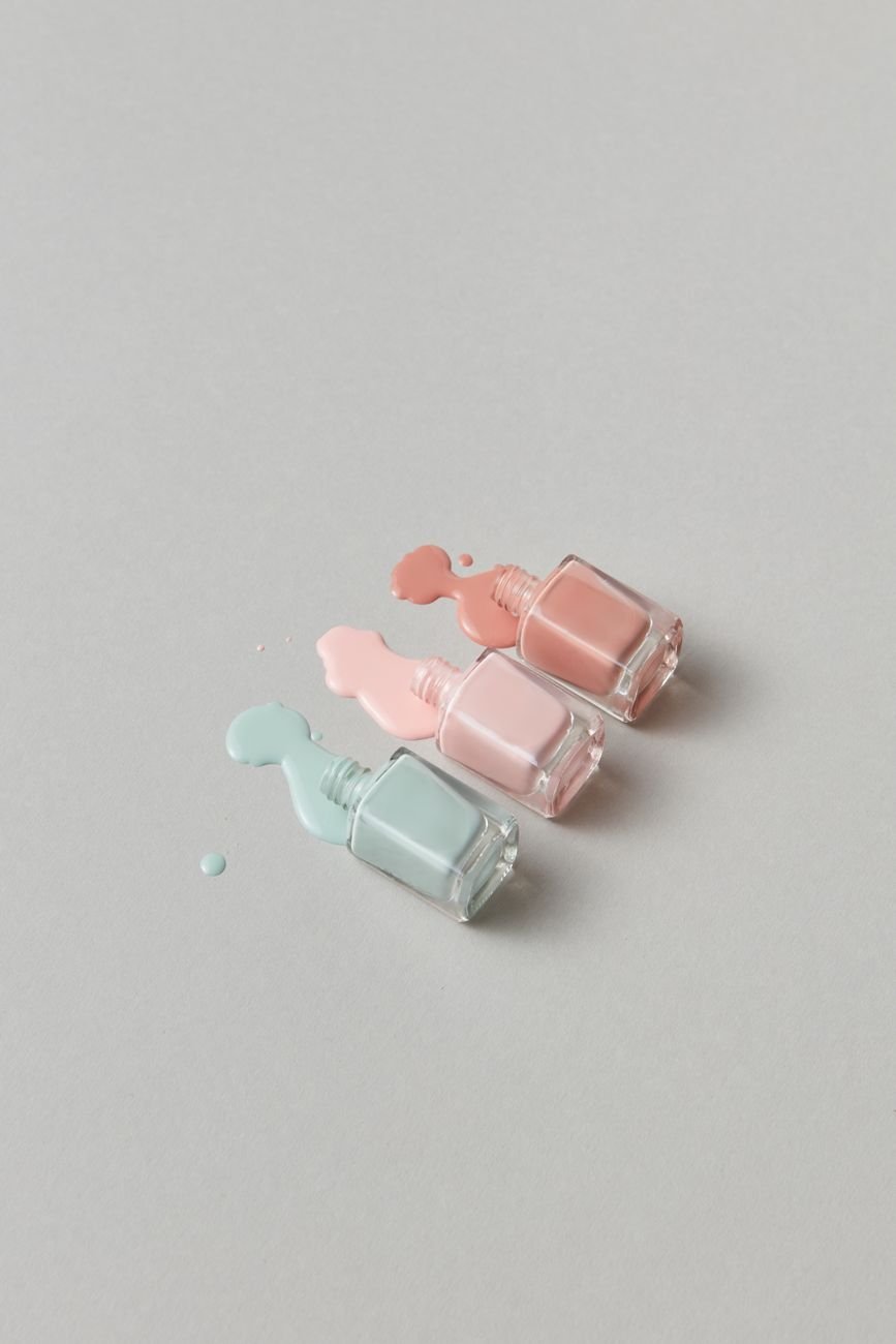 Three bottles of pastel nail polish artfully arranged — soft rose, peach, and lilac.